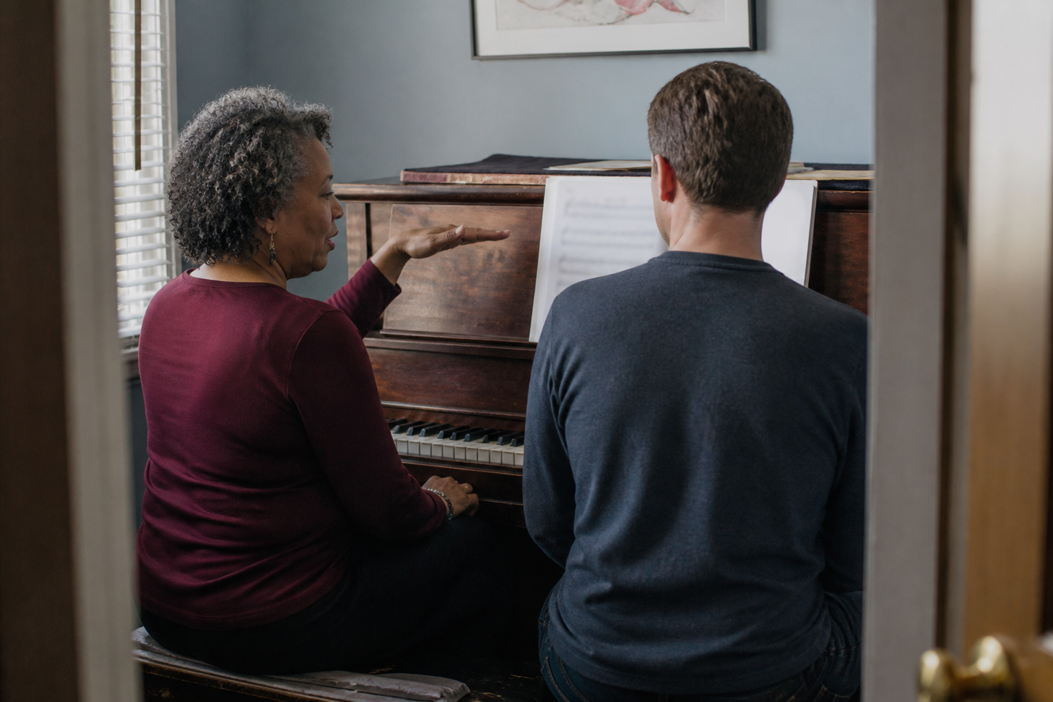 Piano teacher sitting beside adult student at piano, pointing to sheet music and guiding hand position